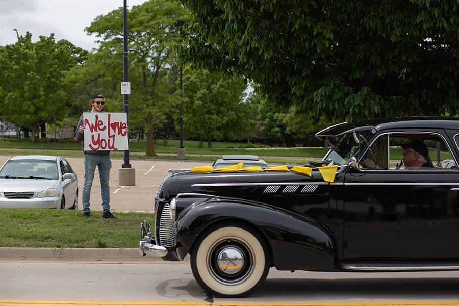 Dean Rodney Miller drives a classic car through campus, and a young man standing on the sidewalk holds a sign that says "We Love You."
