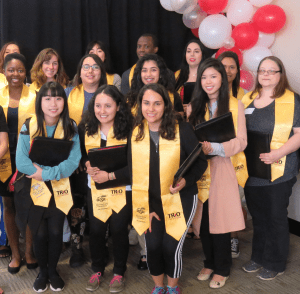 Several TRIO Student Support Services graduates gather for a group photo wearing their TRIO stoles and holding their graduate ceremony gift of a zippered pleather portfolio with big smiles.