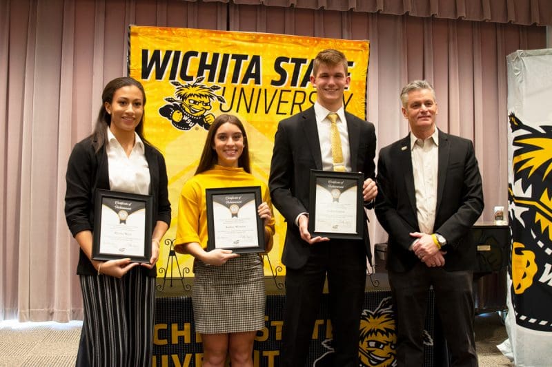 The three winners stand with President Muma holding their certificates behind big WSU banner.
