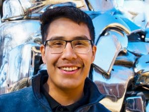 Juan is smiling and standing in front of a chrome statue on campus.