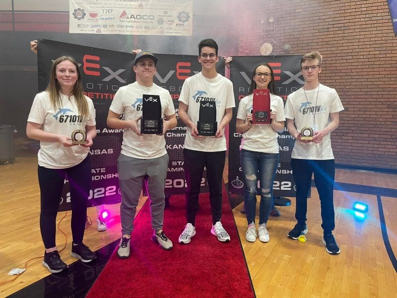 Five white adolescents (two girls and three boys) stand side-by-side and a red carpet holding metal trophies of varying sizes and shapes. They are wearing matching white t-shirts with a team logo that reads "67101V" and has a blue dolphin jumping over the text. In the background are three black banners each with the words "VEX Robotics Competition Kansas State Championship 2022".