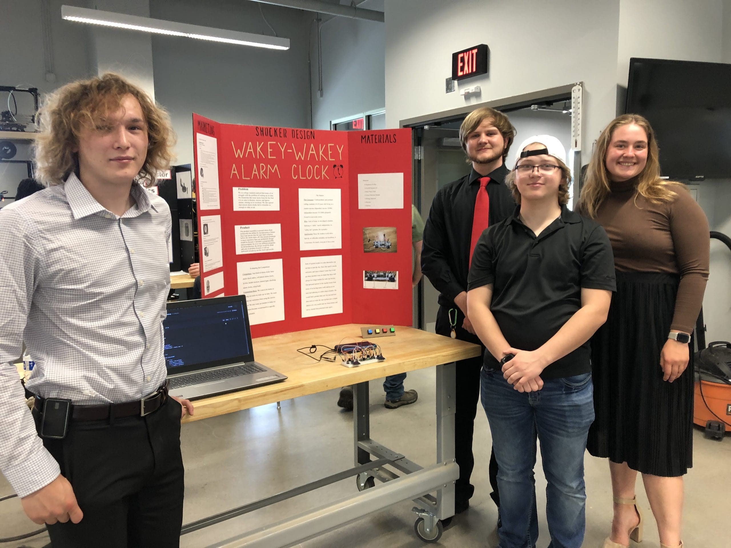 A group of four students smiling at the camera and dressed up to give an open house presentation of their alarm clock prototype. Their tri-fold board is red and titled Wakey Wakey Alarm Clock and details their desigin process.