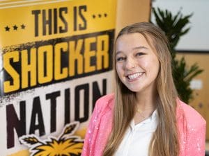 Photo of Maley Hansen in front of a WSU poster that says "This is Shocker Nation"