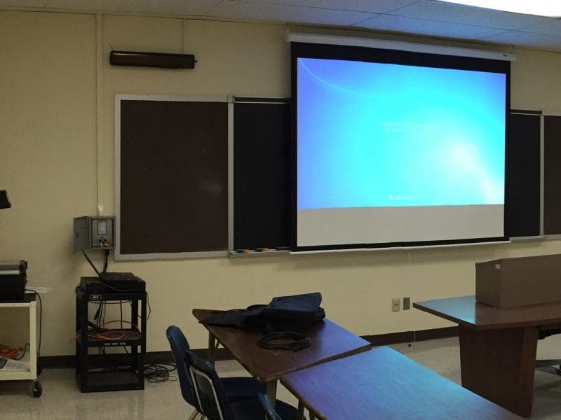 Photo of a classroom with a gray control box on the wall, as well as a transparency projector and projection screen.