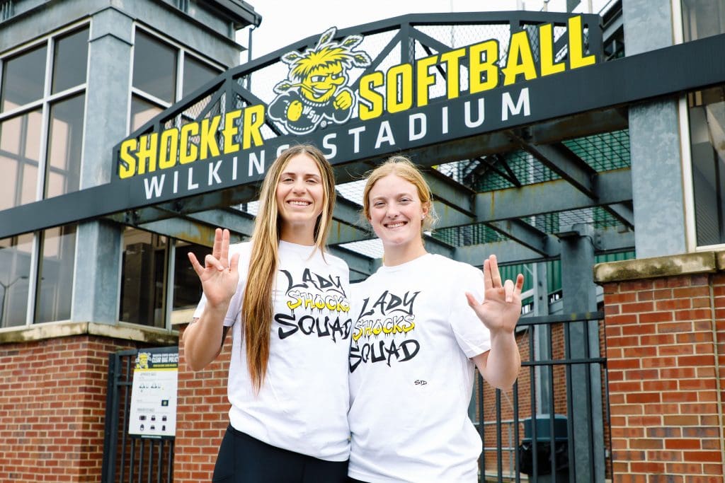 Photo of two students wearing the Lady Shocks Squad shirt while doing the WSU Shockers hand gesture.