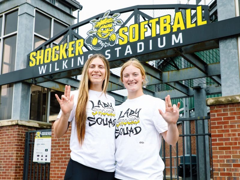 Photo of two students wearing the Lady Shocks Squad shirt while doing the WSU Shockers hand gesture.