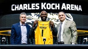 A photo of Terry Nooner holding up a Shocker jersey with President Rick Muma and Kevin Saal, director of athletics inside Charles Koch Arena.