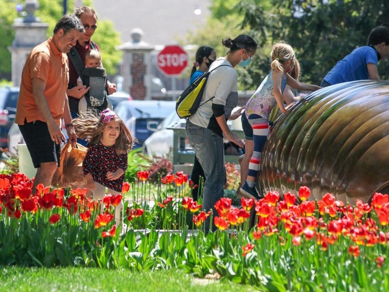 A photo of several family members exploring a large bronze sculpture of a millipede by artist Tom Otterness near the Ulrich Museum outside on a sunny day.