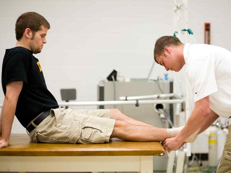 Photo of an athletic training major wrapping the ankle of another student sitting on a taping table platform.