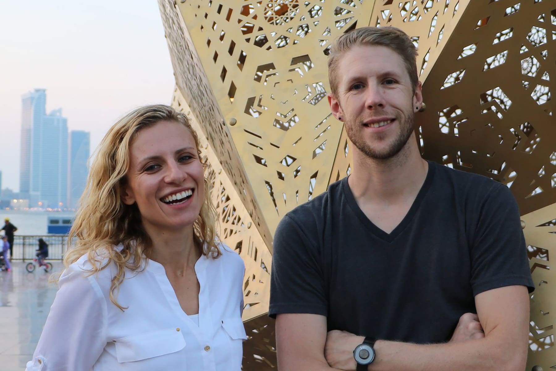 A photo of Yelena Filipchuk and Serge Beaulieu, from the art collective HYBYCOZO, standing in front of a many-sided, brass, geometric sculpture.