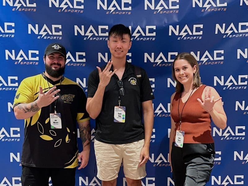 Joe Mazzara, Assistant Director of Esports; Travis Yang, Director of Esports; and Avery Pierce, Assistant Director of Programming; posing in front of the NACE backdrop