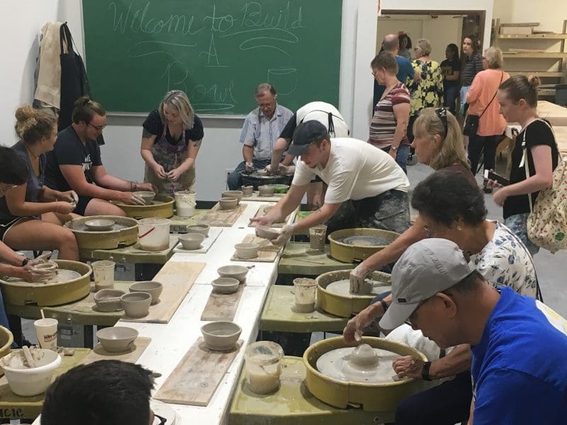 A group of people throw and handbuild ceramic bowls around a table. A chalkboard says, "Welcome to Build A Bowl."