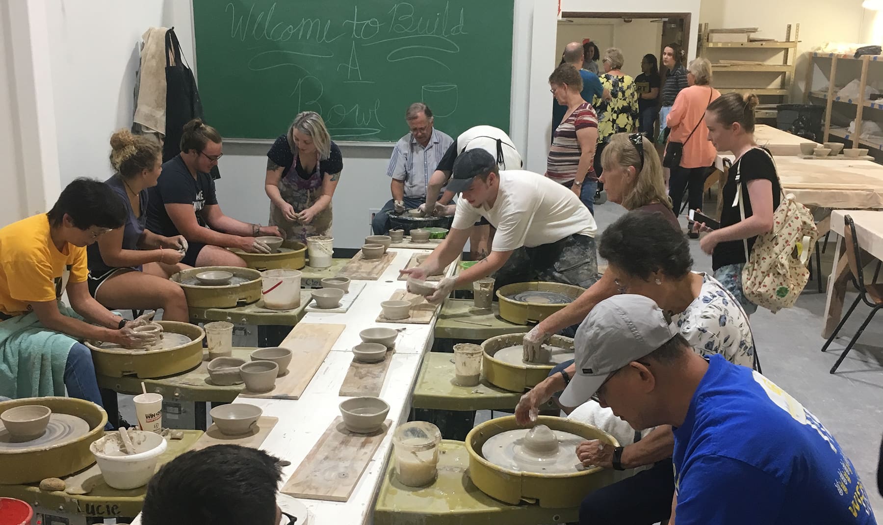 A group of people throw and handbuild ceramic bowls around a table. A chalkboard says, "Welcome to Build A Bowl."