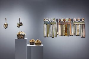 Ceramic works that look like baskets of cotton sit on two pedestals in front of six hanging ceramic works on a wall.
