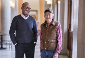 A middle-aged man and an older man pose indoors before some columns.