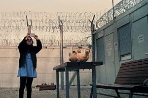 A girl in hijab takes a photo in an outdoor space surrounded by razor wire. A teddy bear sits nearby.