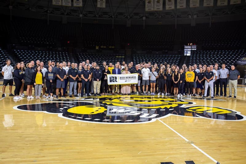 President Rick Muma holds a Rick Rewards sign with members of the Athletics Department in Charles Koch Arena