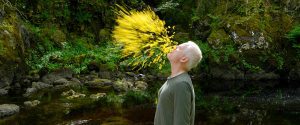 A man with white hair blows yellow flower petals into the air as he stands in front of a pond and moss covered bolders.