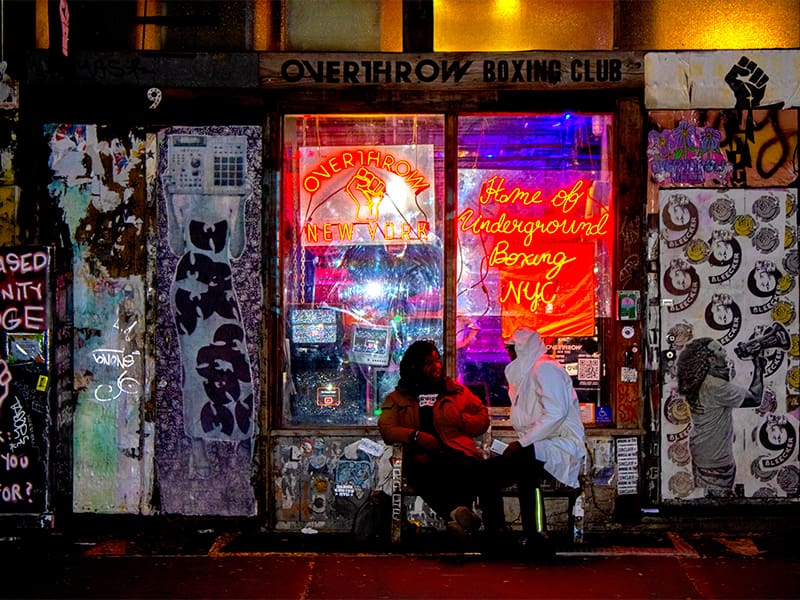 Two figures sitting on a bench in front of Overthrow Boxing Club in New York City. The building is completely covered by posters, graffiti, and miscellaneous destressed signage. The phrases "Overthrow New York" and "Home of Underground Boxing NYC" are displayed via neon signs above the sitting figures. It is night.