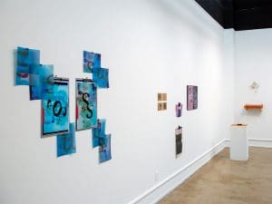 An art gallery with white walls and various artworks, including abstract collages, prints, and clipboards with colorful designs.