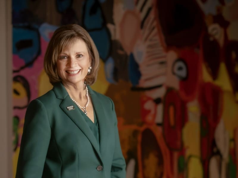 Elizabeth King poses in a hunter green blazer with a WSU pin in front of vibrant artwork.