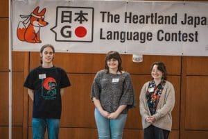 Image shows WSU student Eve Moore standing on stage with the other two winners receiving her prize below a banner that reads "Heartland Japanese Language Contest."