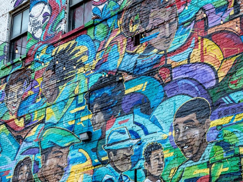 A mural by one of the Urban Canvas artists, Quintis Pinkston, is painted in spray paint and features smiling people.