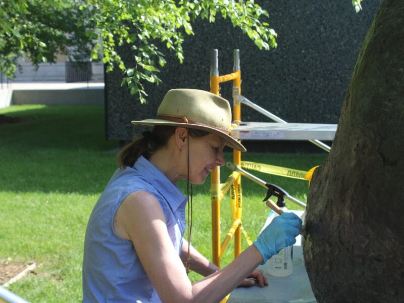 A woman in a hat cleans a sculpture with a dry paintbrush.