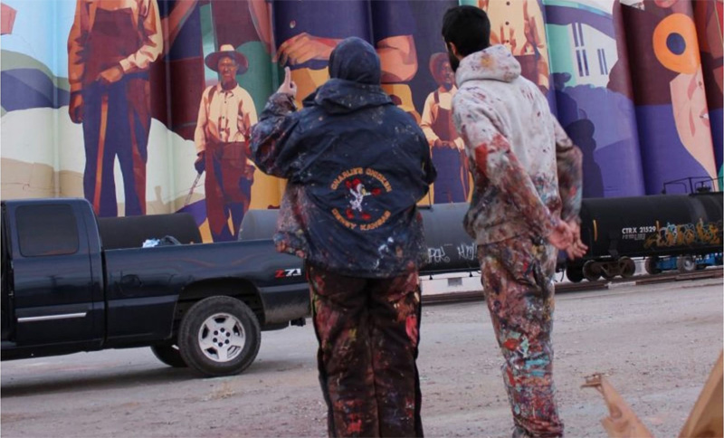 Two artists dressed in painted clothing discuss work they are doing on a large mural.