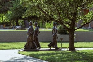 Sculptures of three women walking are located under a leafy tree.
