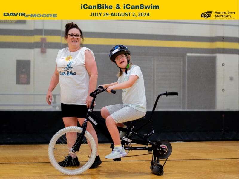 The heading reads iCanBike and iCanSwim and the subheading reads July 29 - August 2, 2024. The Davis Moore and Wichita State University Department of Physical Therapy logos are also included in the heading in front of a yellow background. The picture is of two women. One is a young lady on a specialized bicycle wearing a helmet who has paused while riding the the bike to smile for the camera. The other is of a smiling woman who is has paused mid-step. She is wearing a t-shirt with sleeves rolled up that reads iCanBike with a bicycle on it.