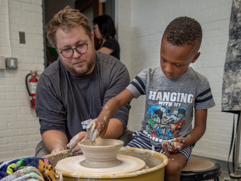 A boy throws a ceramic bowl with the help of a man.