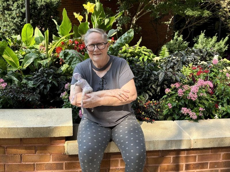 An artist poses with a handmade felted figure in a garden.