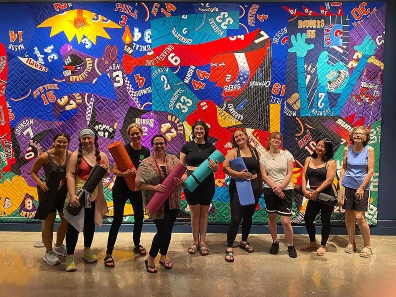 A group of yoga practitioners pose with their yoga mats in front of a large art quilt.