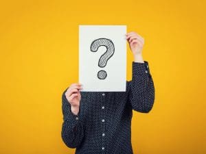This is a person in a black shirt holding a sign covering their face with a question mark on it. The background is a gold color.
