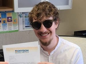 Photograph of speaker, Jeremy Barton, smiling at the camera and holding a piece of paper that has the Envision Research Institute letterhead. Jeremy is wearing round sunglasses.