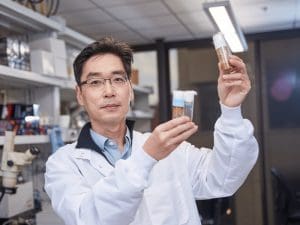Photograph of Dr. Wan Hee Yoon in a research lab. He is slightly smiling at the camera, while holding two tubes of biological samples in each hand.