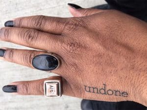 A hand with long nails painted black and a tattoo that says, "undone."