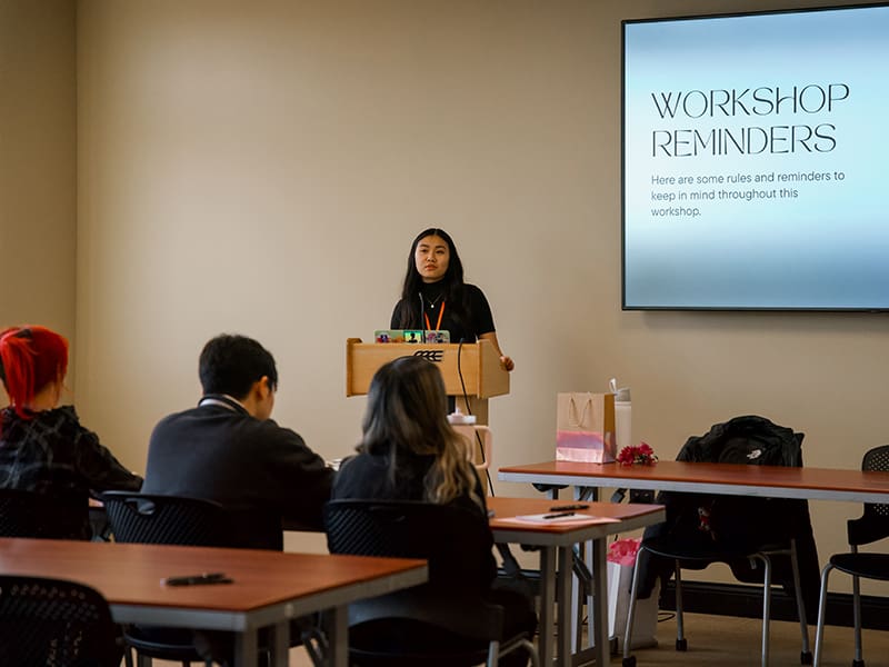 Donna Tran speaks to a group at a workshop. The screen behind her reads, "Workshop Reminders, here are some rules and reminders to keep in mind throughout this workshop."