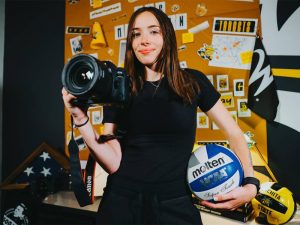 Skylar Fleeman, a WSU grad student who works in Shocker Athletics, poses with a camera and volleyball in front of a wall of Shocker Athletics gear