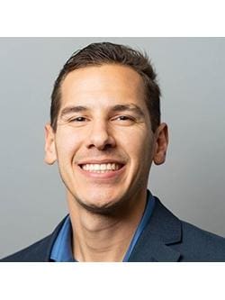 The image is a professional headshot of Samuel "Sam" Jones. He is smiling warmly, wearing a dark blue blazer over a medium blue collared shirt, with a neutral gray background. His expression is confident and approachable.
