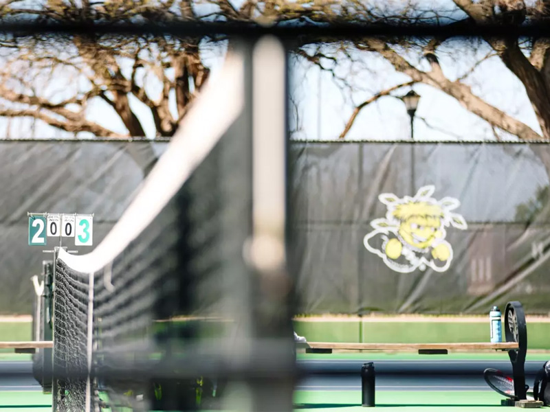 A profile shot of a tennis court net with benches and the WuShocker logo in the background. A board shows the score is 2 to 3.
