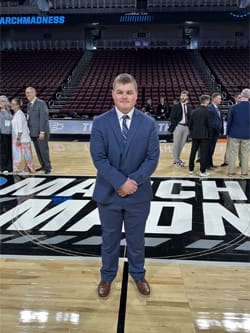 WSU student Liam Crawford stands on the court at the NCAA tournament venue in a navy suit and striped tie. The March Madness logo is visible beneath his feet, with event staff and attendees in the background.