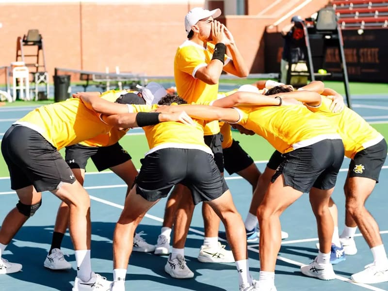 Men's tennis players form a ring around another play yelling in preparation of a game.