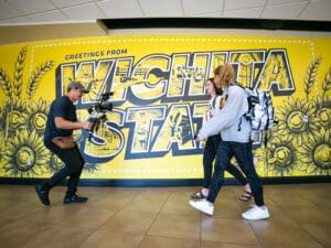 Students walking in front of the Wichita State mural in the Rhatigan Student Center as they're filmed by a cameraman for a TV show.
