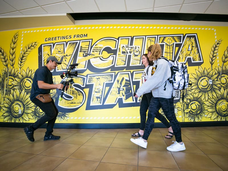Students walking in front of the Wichita State mural in the Rhatigan Student Center as they're filmed by a cameraman for a TV show.