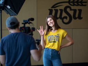 A student poses in front a camera for the filming of a TV show. A black WSU sign is in the background.