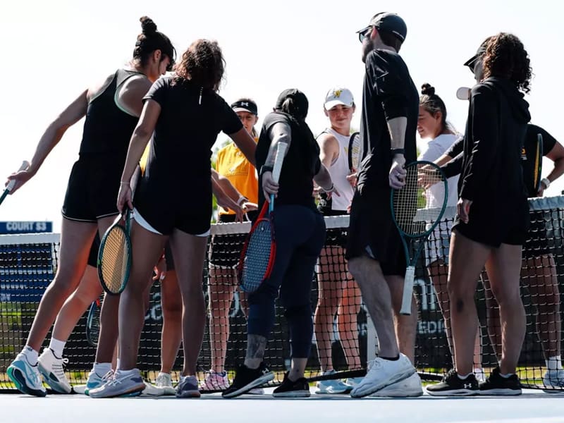 The women's tennis team huddles over a net during a game.