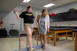 A student in a Wichita State University T-shirt practices a balance and movement assessment with a measurement tool while an instructor watches and provides guidance in the Athletic Training Lab. The lab features exam tables, anatomical models, and clinical equipment.