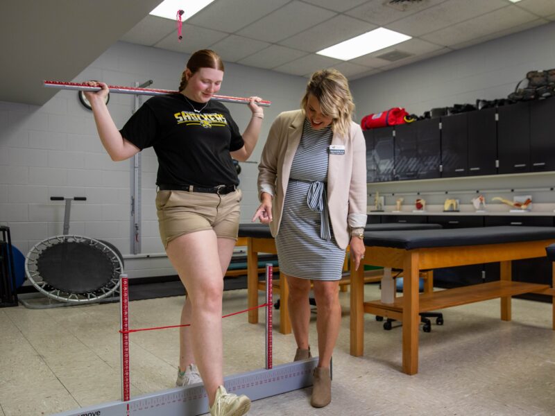 A student in a Wichita State University T-shirt practices a balance and movement assessment with a measurement tool while an instructor watches and provides guidance in the Athletic Training Lab. The lab features exam tables, anatomical models, and clinical equipment.